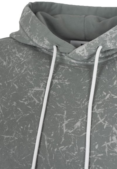 Bluza z kapturem w jasnoszarym materiale z teksturowanym białym abstrakcyjnym wzorem. Posiada białe sznurki ściągające oraz miękki, zamknięty kaptur.