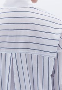 Camisa blanca con rayas verticales azules, que presenta un collar doblado y un diseño en capas en la parte posterior con costuras para dar forma.