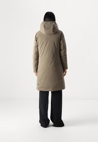 Lang, olivengrøn isoleret frakke med hætte og sømdetaljer, kombineret med mørke bukser med brede ben. Glat tekstur og en afslappet silhuet.