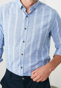 Camicia azzurra chiaro con righe bianche verticali, maniche corte e bottoni marroni. Realizzata in un tessuto testurizzato, adatta per un abbigliamento casual.
