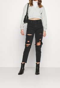 Jeans noirs déchirés avec des bords effilochés, associés à un sweat-shirt court gris clair et des bottes à talons noires. Un petit sac à bandoulière noir est porté.