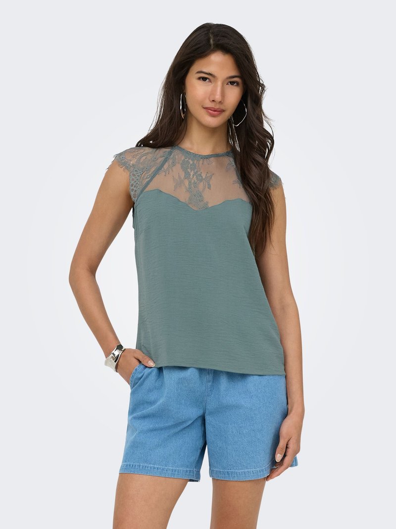 Femme portant un haut en dentelle verte sans manches et un short en jean bleu clair, debout avec une main dans la poche devant un fond uni.