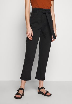 Persona vistiendo pantalones negros de tiro alto con cinturón de tela atado, puños remangados, una blusa naranja quemado y sandalias negras de punta abierta, de pie contra una pared blanca.