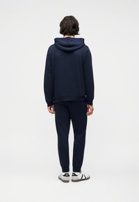 Άνδρας με σκούρα κυματιστά μαλλιά γυρισμένος πλάτη, φοράει μπλε σκούρα φούτερ με κουκούλα, παντελόνι φόρμας σε ταιριαστό χρώμα, λευκά αθλητικά παπούτσια με μαύρες λεπτομέρειες, στέκεται σε απλό φωτεινό φόντο.