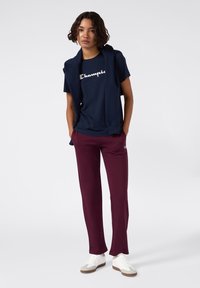 Granatowa bawełniana koszulka z białym logo "Champion", w połączeniu z burgundowymi spodniami dresowymi i białymi sneakersami. Model nosi granatowy sweter zarzucony na ramionach.