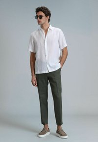 Chemise blanche en lin à manches courtes, pantalon tapered vert olive, et chaussures à enfiler brun clair. Le modèle porte des lunettes de soleil noires, se tenant contre un fond gris.