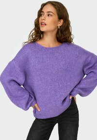 Maglione lavorato a maglia oversize di colore viola con un design testurizzato a costine e maniche ampie, abbinato a jeans neri. Tessuto morbido e accogliente.