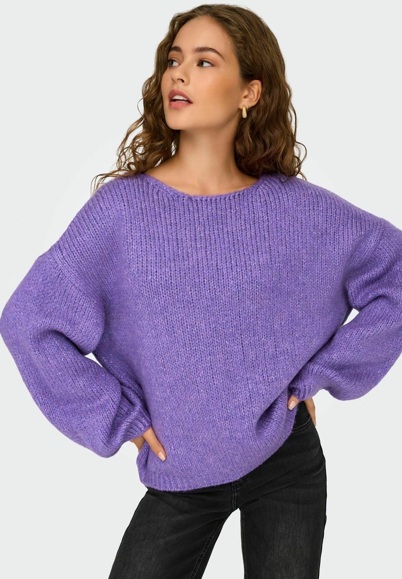 Maglione lavorato a maglia oversize di colore viola con un design testurizzato a costine e maniche ampie, abbinato a jeans neri. Tessuto morbido e accogliente.