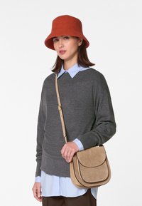 Grauer Strickpullover, hellblaues Hemd mit Kragen, braune Hose, braune Umhängetasche und ein orangefarbener Filzhut.