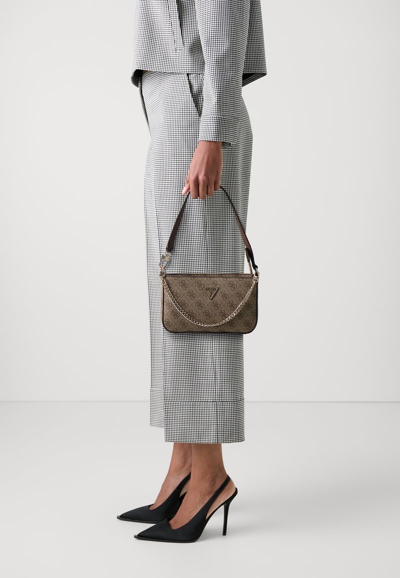 Personne portant un pantalon et un blazer à carreaux noir et blanc, tenant un sac à main marron à motif avec un détail chaîne, portant des talons noirs.