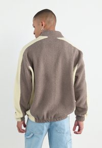 Bruine en crèmekleurige fleece jas met een hoge kraag, elastische zoom en lange mouwen. Beschikt over een textuur oppervlak en contrasterende kleurpanelen.