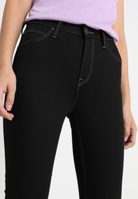 Los jeans de mezclilla negra presentan costuras blancas en contraste, un cierre de botón y un diseño ajustado, con una textura suave y el clásico estilo de cinco bolsillos.