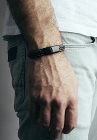 Brățară din piele împletită de culoare maro, cu un închizător din metal negru, purtată pe o mână pe un fundal gri deschis. Design texturat și rezistent.