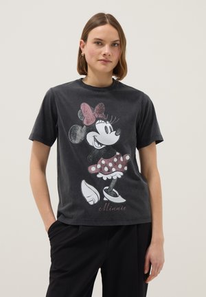 Femme aux cheveux bruns et raides portant un t-shirt noir arborant Minnie Mouse vintage avec un nœud et une jupe à pois, accompagnée d'un pantalon noir.