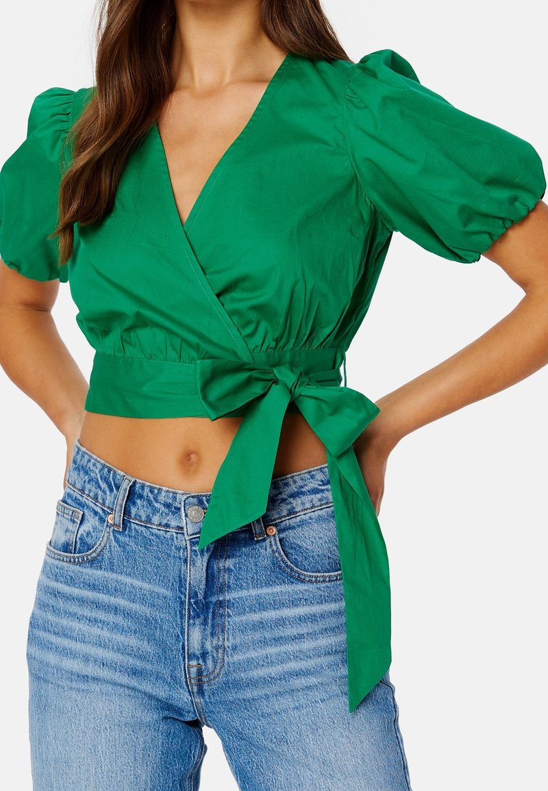 Top verde con maniche a sbuffo, cintura annodata e scollatura a V profonda. Abbinato a jeans in denim azzurro chiaro a vita alta, che mostrano una texture liscia.