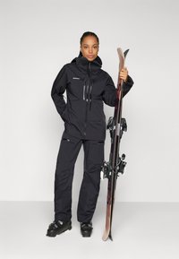 Zwarte skijack en -broeken met ritsen en zakken, gecombineerd met zwarte skibotten. Persoon houdt ski's in één hand.