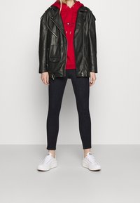 Svart läderbikerjacka, röd hoodie med dragsko, mörka slim-fit jeans och vita sneakers. Jackan har dragkedjor och fickor.