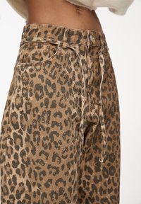 Pantalon à imprimé léopard marron avec une coupe taille haute, doté de poches avant et d'un cordon de serrage. Le matériau semble texturé et doux.