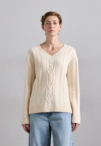 Pull en maille torsadée couleur crème avec un col en V, présentant des motifs tressés raffinés et des poignets et un ourlet côtelés. Associé à un jean bleu clair.