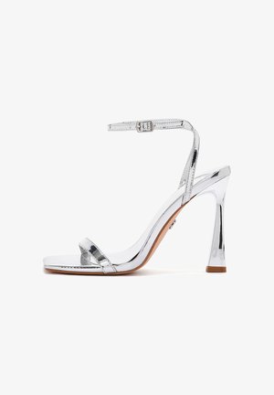 Zilveren sandaal met hoge hak, dunne enkelriem met gesp, open teen en blokhak op een witte achtergrond.