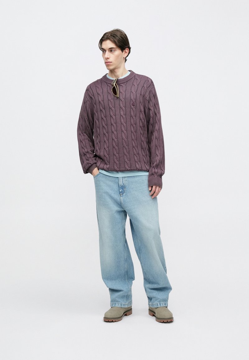 Gestrickter lila Pullover mit Zopfmuster, hellblauer Kragen, getragen über einem türkisfarbenen Hemd. Hellblaue Jeanshose und taupefarbene Stiefeletten.