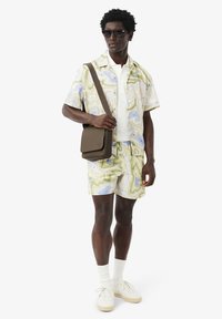 Completo con camicia a maniche corte e pantaloncini in fantasia marmorizzata verde pastello e blu, abbinato a t-shirt bianca, sneakers color crema e borsa a tracolla marrone.