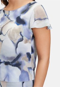 Leichte Bluse mit fließendem Design, die ein blaues und graues abstraktes Muster aufweist, kurze Schmetterlingsärmel und einen runden Halsauschnitt hat.