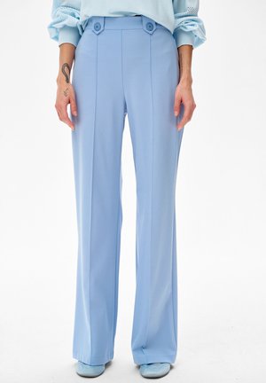 Persoon draagt lichtblauwe, hoog uitgesneden broek met wijde pijpen en knoopdetails, gecombineerd met bijpassende schoenen, staand tegen een effen witte achtergrond.