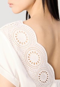 Top blanc doté d'un dos en forme de vagues avec des broderies en œillets détaillées et des découpes circulaires. Tissu doux avec des bords de manches retournés.
