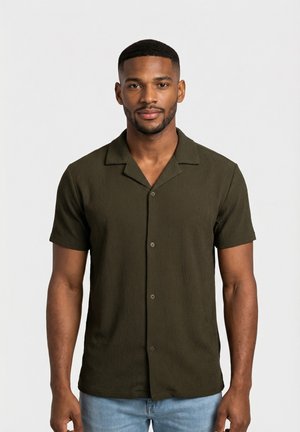 Uomo con capelli corti e barba curata che indossa una camicia verde scuro a maniche corte con bottoni e jeans azzurri, in piedi contro uno sfondo semplice.