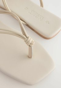 Beiga flip-flops med fyrkantig tå, med tunna, knutna remmar och en slät sula. Sula har en ljusbrun accent.