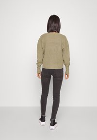 Olivově zelený pletený svetr s dlouhými rukávy, nošený přes černé skinny džíny a černé vysoké tenisky, viděný zezadu.