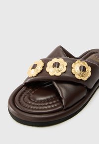 Brown leather slide sandal with cross-strap design, three ornate gold floral embellishments, and a cushioned insole with stitching detail.