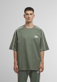 Olijfgroen katoenen t-shirt met korte mouwen, ronde hals en witte zijaccenten. Beschikt over een logopatch op de borst. Ontspannen pasvorm.