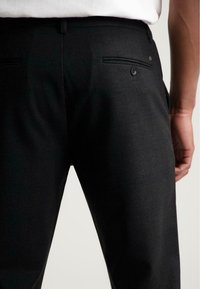 Schwarze maßgeschneiderte Hose mit glatter Textur, ausgestattet mit Seitentaschen und einer Gesäßtasche mit einem Knopfdetail.