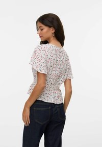Kvinna med långt mörkt hår som bär hoopsörhängen, en vit blommig kortärmad blus med smockad midja och mörkblå jeans, vänd mot vänster.