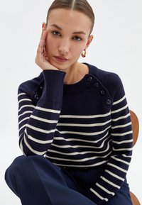 Marineblau gestreifter Pullover mit weißen Streifen und geripptem Kragen, mit vier Knopfakzenten an der Schulter und strukturiertem Strickstoff.
