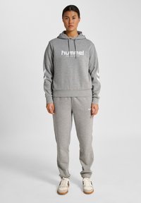 Vrouw staand in een grijze hummel hoodie en bijpassende trainingsbroek met witte sneakers tegen een effen lichte achtergrond.