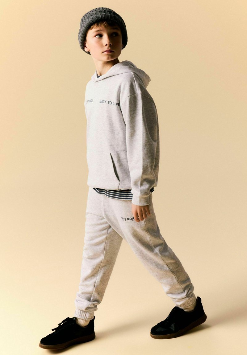 Jeune garçon en sweat à capuche gris clair et pantalon de survêtement, portant un bonnet gris foncé et des baskets noires sur fond beige.