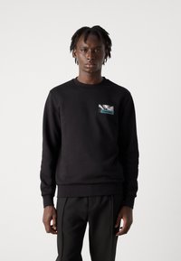 Zwarte sweatshirt met ronde hals en lange mouwen. Heeft een kleine voorkeurszak met een grafisch ontwerp. Gemaakt van een zachte, textuurachtige stof.
