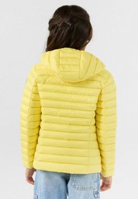 Geel gewatteerd jack met capuchon, voorzien van horizontale doorgestikte segmenten en een gladde textuur, gecombineerd met lichtblauwe spijkerbroek.