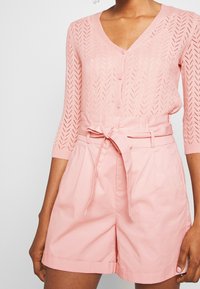 Cardigan court en maille rose pâle à manches courtes, col en V et fermeture à boutons, assorti à un short taille haute rose avec une ceinture nouée et des poches.