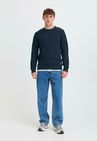 Dark blue ribbed knit sweater with a round neckline, layered over a light gray shirt, paired with loose blue denim jeans and gray sneakers.
