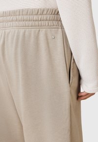 Pantalones de chándal beige con una cinturilla elástica ancha y bolsillos laterales, con una textura suave y un pequeño detalle metálico.