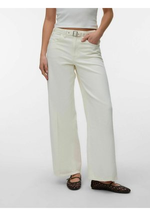 Femme portant un pantalon blanc taille haute à jambes larges avec ceinture et des ballerines tissées noires, debout avec une main dans la poche sur fond blanc.