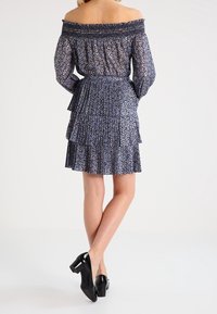 Vestido azul marino off-the-shoulder con escote fruncido, mangas largas, tejido texturizado y dobladillo con volantes en capas. Combinado con zapatos de tacón bloque negro.