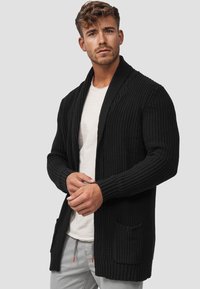Cárdigan negro de punto con textura de canalé, de frente abierto, mangas largas y dos bolsillos frontales. Llevado sobre una camiseta clara y pantalones grises.