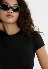 Schwarzes, figurbetontes T-Shirt mit kurzen Ärmeln, kombiniert mit eleganten schwarzen Cat-Eye-Sonnenbrillen. Welliges, dunkles Haar und glatte Haut sind sichtbar. Weißer Hintergrund.