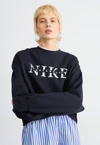 Crna dukserica od pamuka s bijelim Nike logom. Dizajn uključuje duge rukave i okrugli izrez, u kombinaciji s prugastim hlačama.