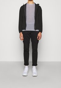 Person, der einen schwarzen Reißverschluss-Hoodie, ein weiß-schwarz gestreiftes T-Shirt, schwarze Cargo-Hosen, weiße Socken und weiße Sneakers trägt.